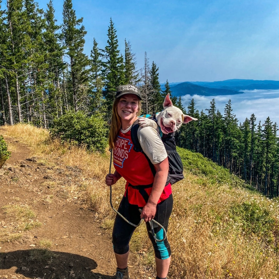Girl wearing a red Rope Hounds shirt hiking with her white pitbull dog. The dog is in a backpack and they are hiking in a tree-covered mountain with water in the distance and blue skies above.